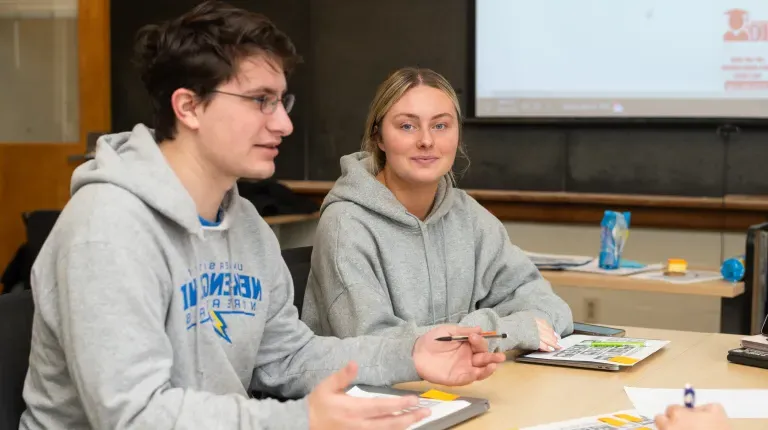 Two students discuss a book in a class with a blackboard behind them